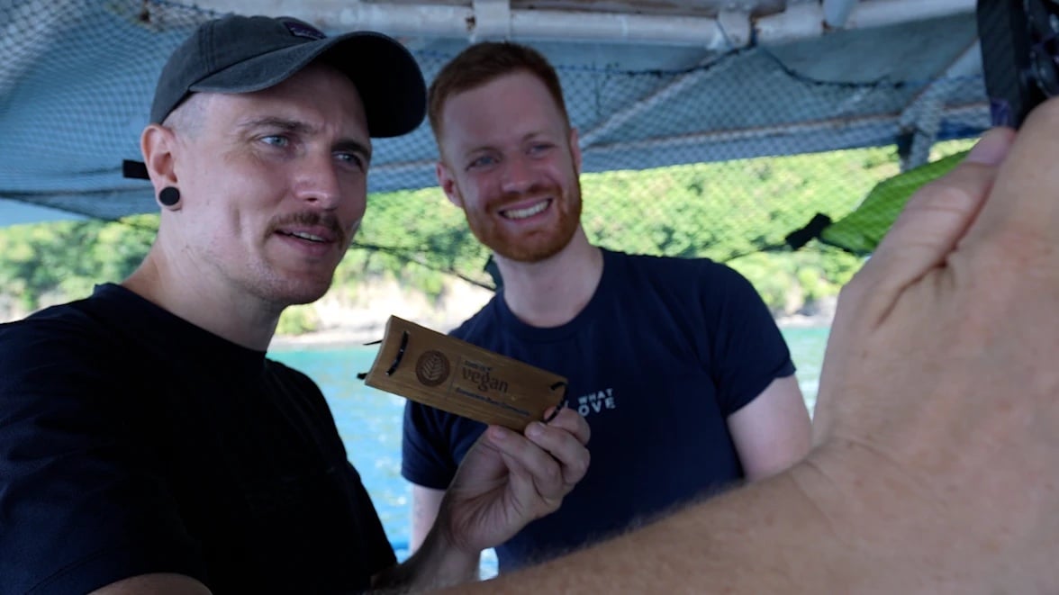 Yannick von This Is Vegan und YouTuber Manuel Bergmann halten auf dem Tauchboot die Holz-Plakette der Community-Korallenspende in den Händen