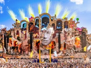 Die Mainstage von Tomorrowland 2026 mit gelben Pyro-Fontänen vor blauem Himmel und riesiger Menschenmenge