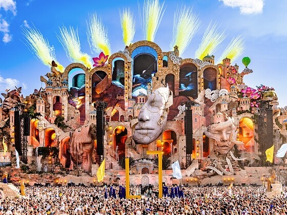 Die Mainstage von Tomorrowland 2026 mit gelben Pyro-Fontänen vor blauem Himmel und riesiger Menschenmenge