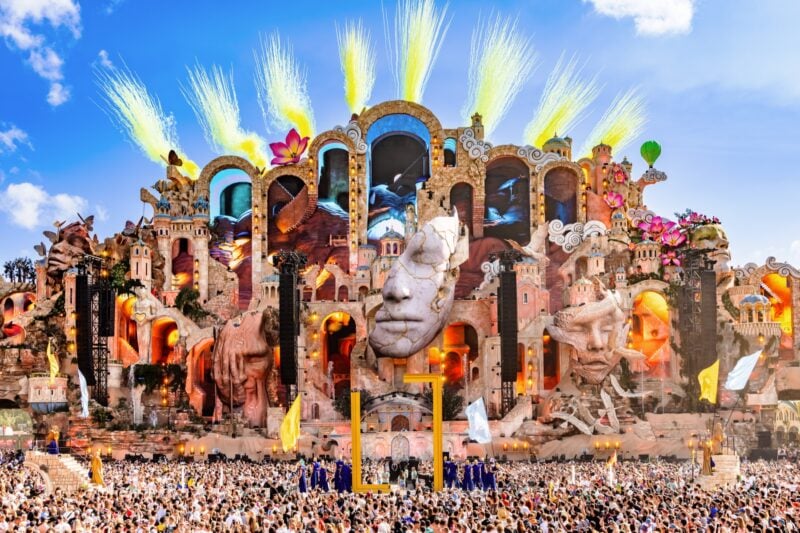 Die Mainstage von Tomorrowland 2026 mit gelben Pyro-Fontänen vor blauem Himmel und riesiger Menschenmenge