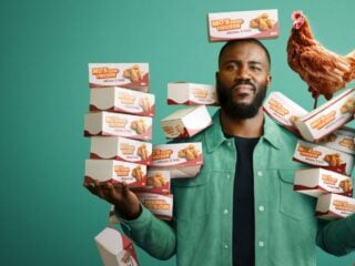 Mo Gilligan steht vor grünem Hintergrund mit Stapeln von Fried-Chicken-Boxen in den Händen, auf seiner Schulter sitzt ein Huhn