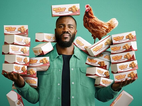 Mo Gilligan steht vor grünem Hintergrund mit Stapeln von Fried-Chicken-Boxen in den Händen, auf seiner Schulter sitzt ein Huhn