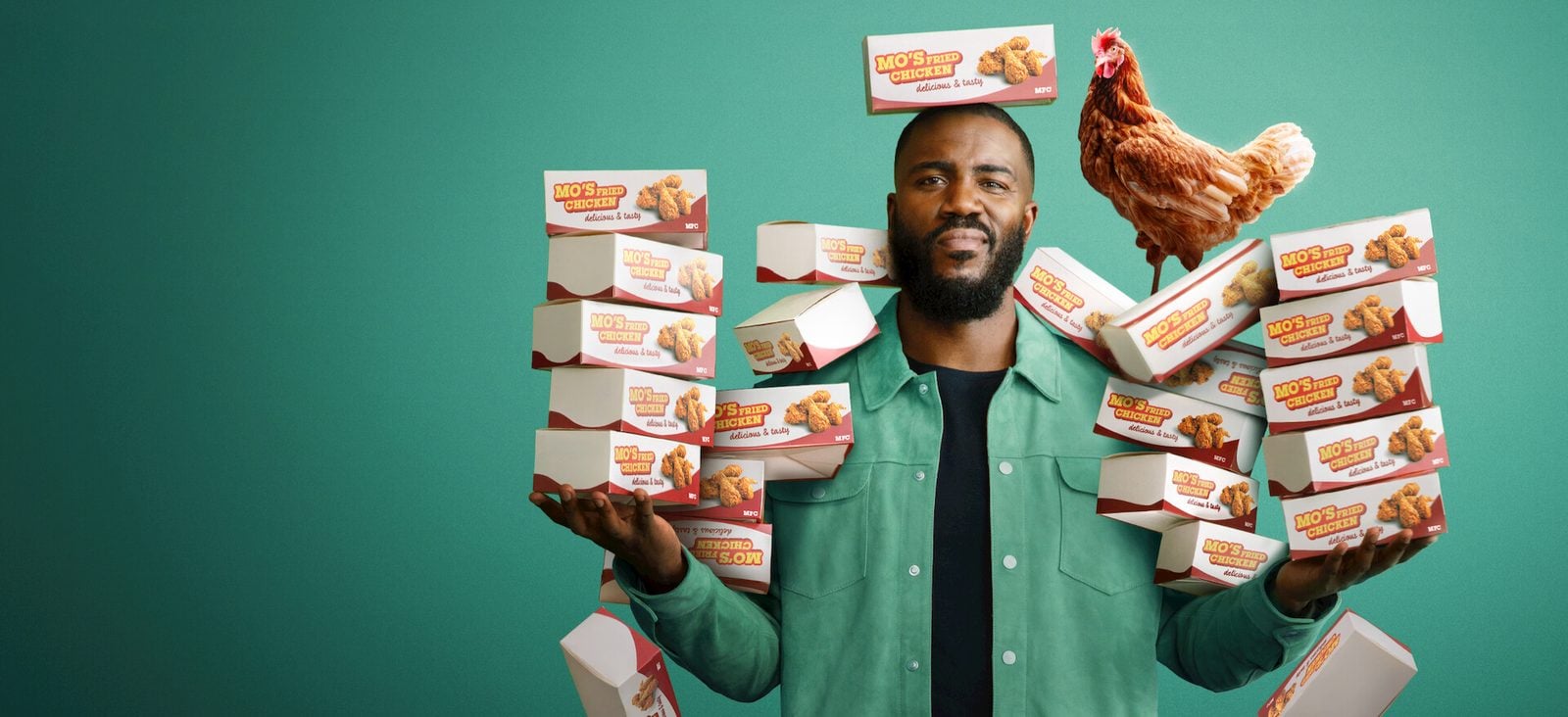 Mo Gilligan steht vor grünem Hintergrund mit Stapeln von Fried-Chicken-Boxen in den Händen, auf seiner Schulter sitzt ein Huhn