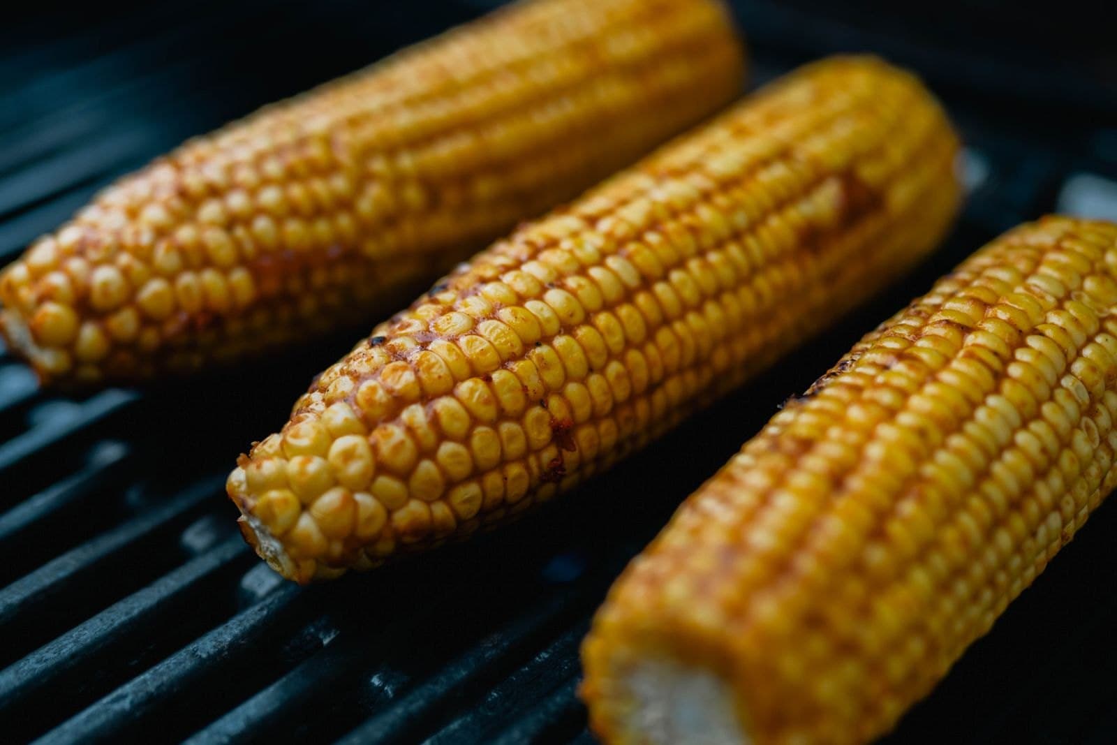 Drei Maiskolben liegen nebeneinander auf einem heißen Grillrost