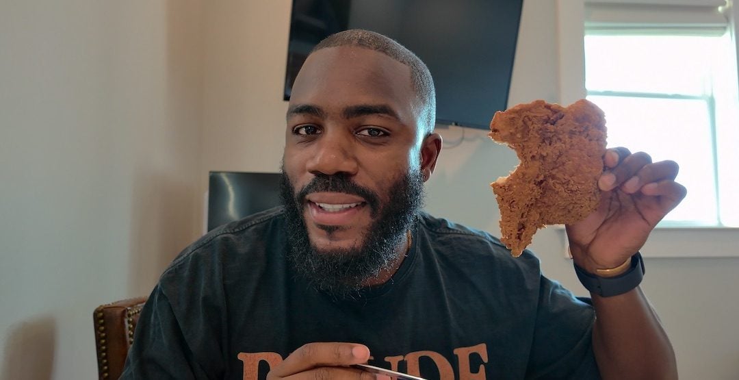 Mo Gilligan sitzt an einem Tisch und hält ein angebissenes Stück frittiertes Hähnchen in die Kamera