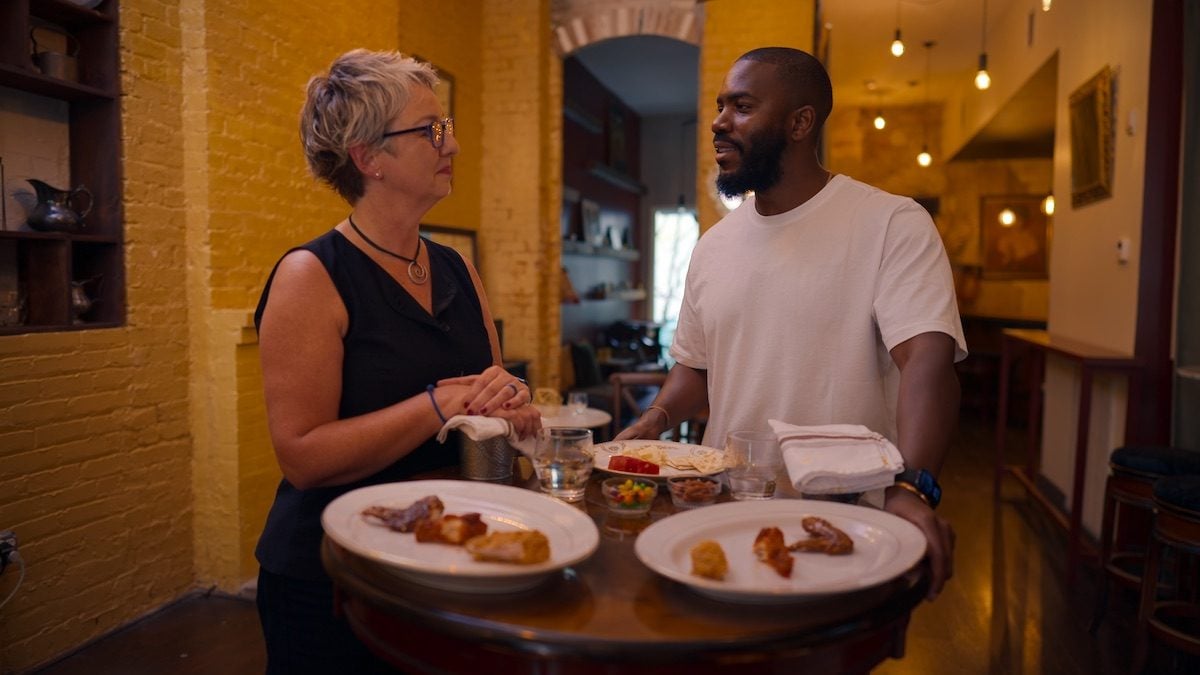 Mo Gilligan steht mit einer Frau an einem runden Tisch in einem Restaurant, auf den Tellern liegen Hähnchenteile