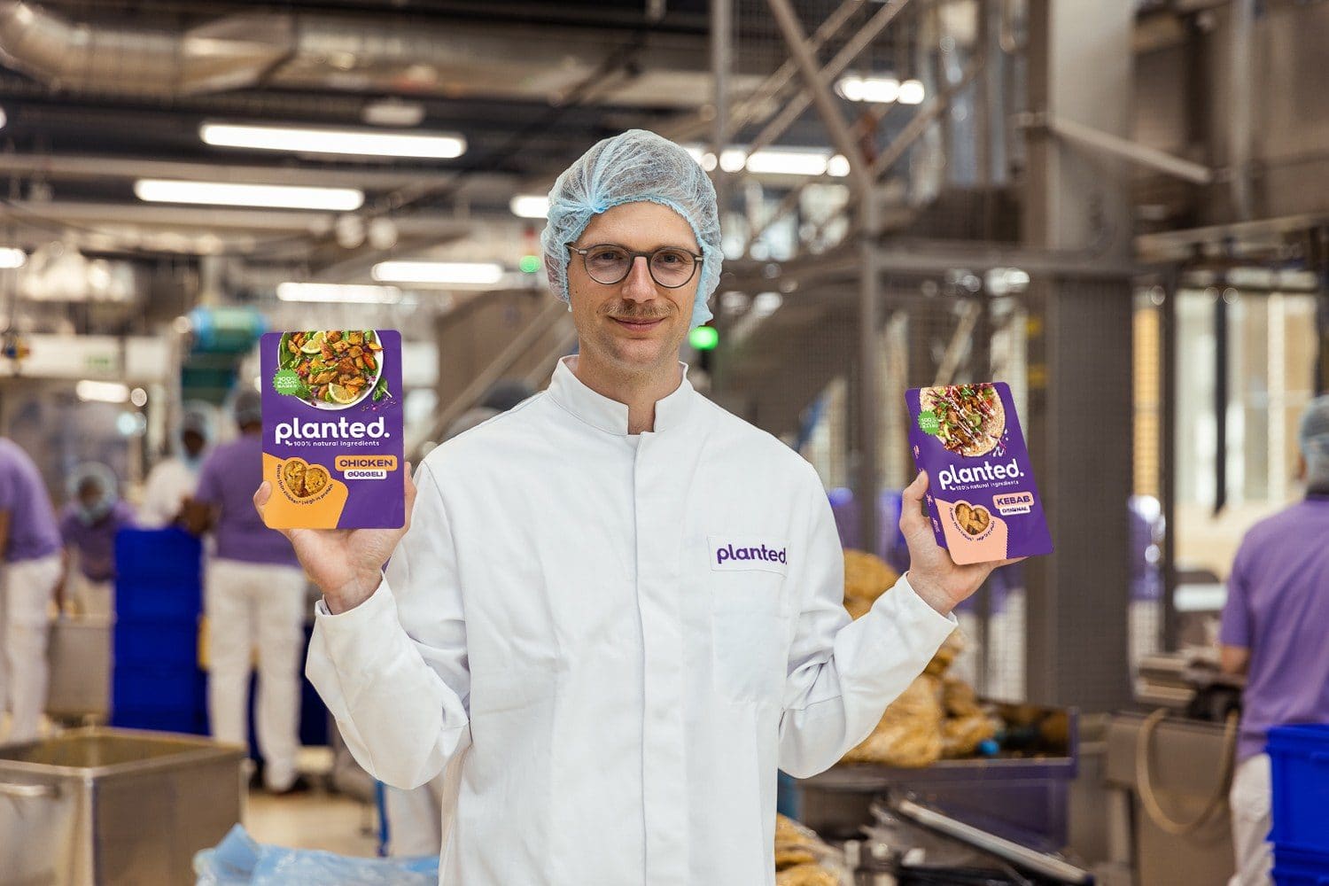 Ein Mitarbeiter in Schutzkleidung hält zwei Packungen Planted-Produkte in der Produktionshalle