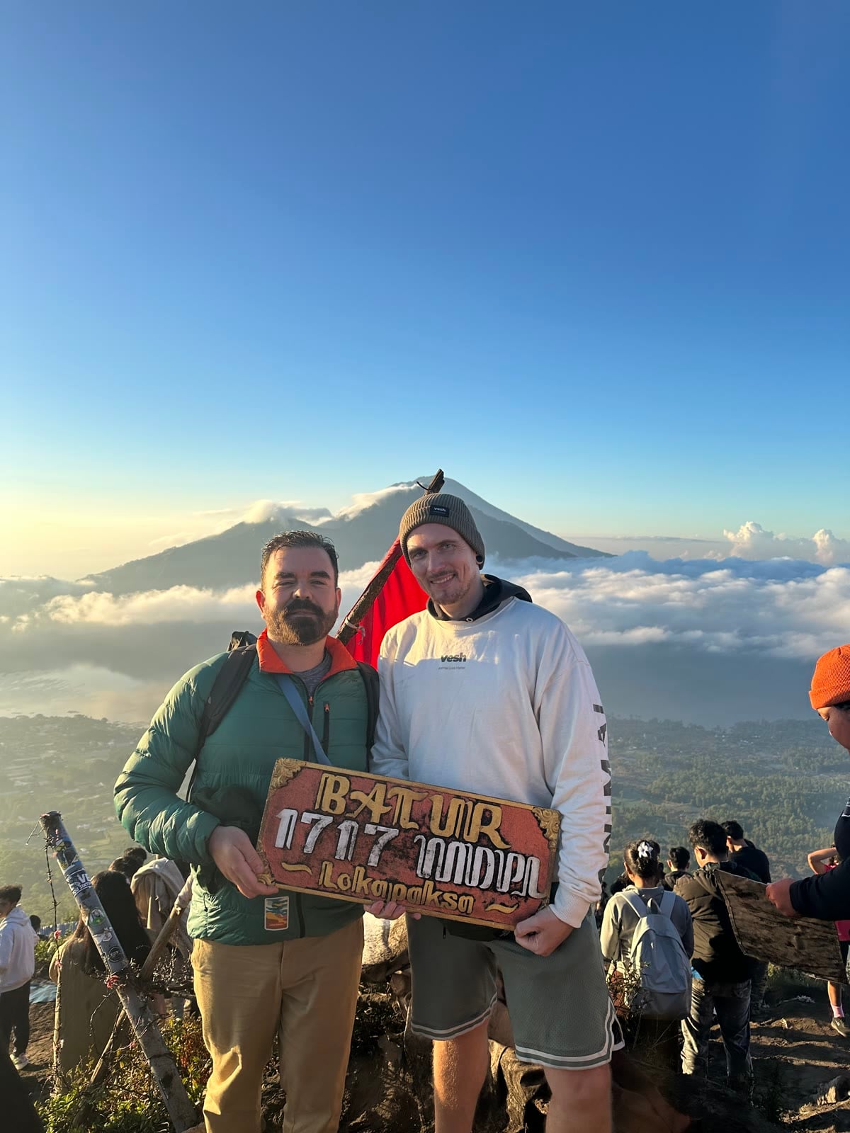 Robbie Lockie und Yannick Haldenwanger bei Sonnenaufgang auf dem Gipfel des Mount Batur auf Bali, mit dem 1717-Meter-Schild und dem Mount Agung über den Wolken im Hintergrund