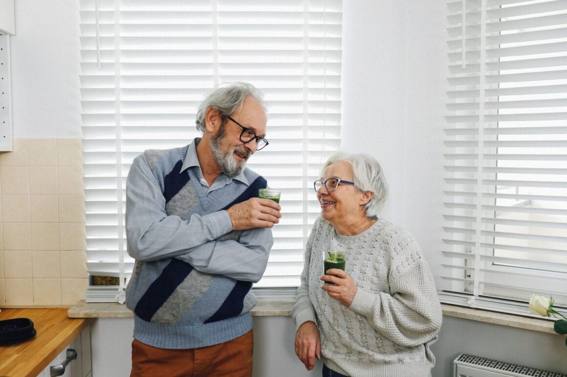 Älteres Paar steht lachend in einer Küche und hält zwei Gläser mit grünen Smoothies
