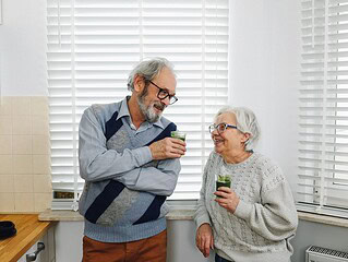 Älteres Paar steht lachend in einer Küche und hält zwei Gläser mit grünen Smoothies