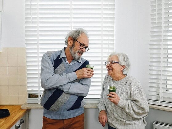 Älteres Paar steht lachend in einer Küche und hält zwei Gläser mit grünen Smoothies