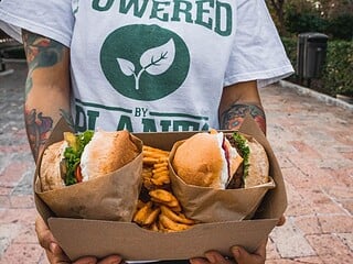 Person in einem Powered-by-Plants-Shirt hält eine Box mit zwei pflanzlichen Burgern und Pommes