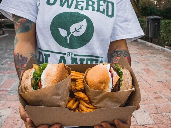 Person in einem Powered-by-Plants-Shirt hält eine Box mit zwei pflanzlichen Burgern und Pommes