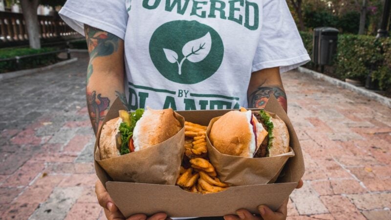 Person in einem Powered-by-Plants-Shirt hält eine Box mit zwei pflanzlichen Burgern und Pommes