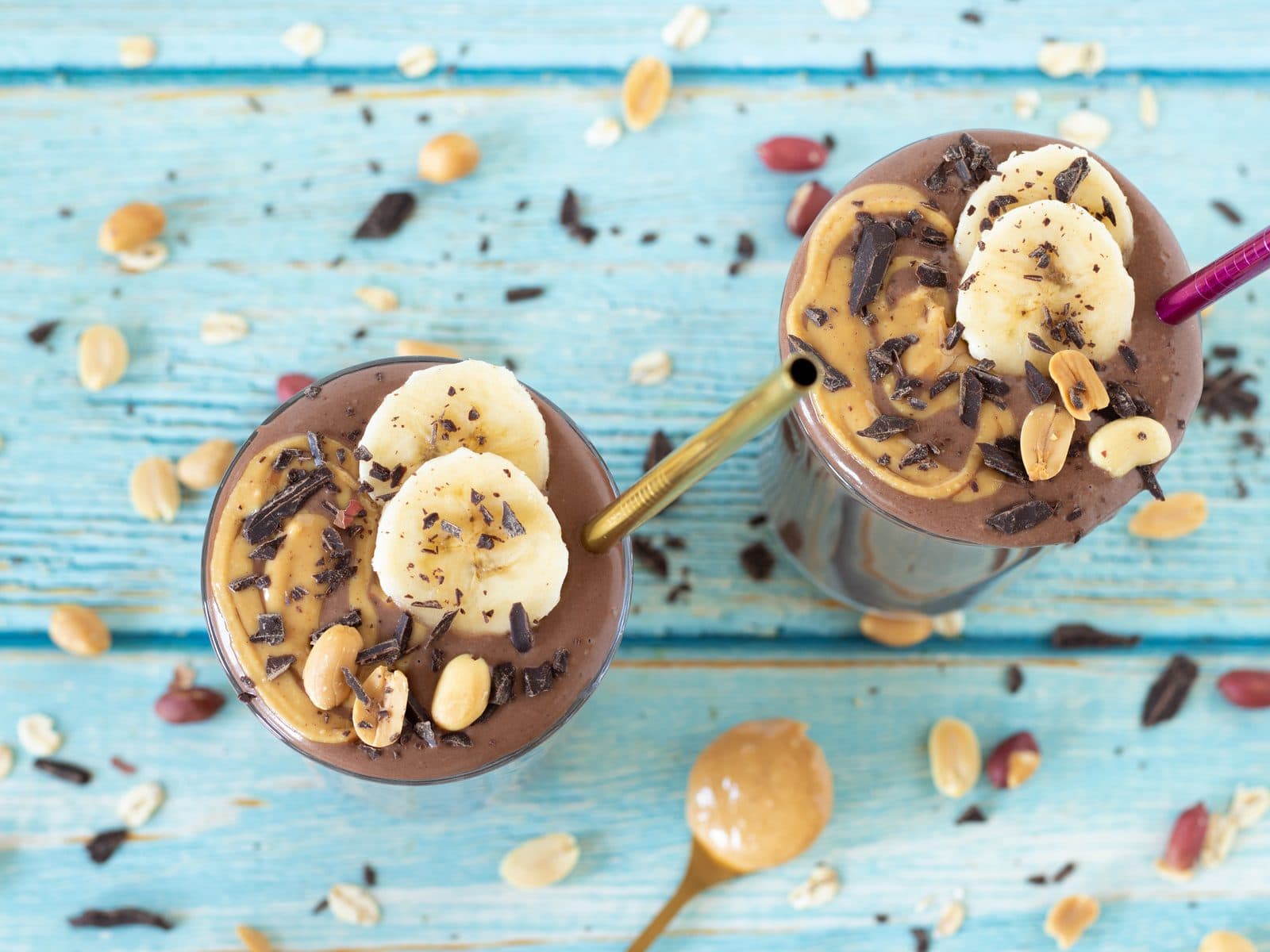 Zwei Schoko-Erdnuss-Bananen-Smoothies mit Bananenscheiben, Schokoraspeln und Erdnuessen auf blauem Holztisch