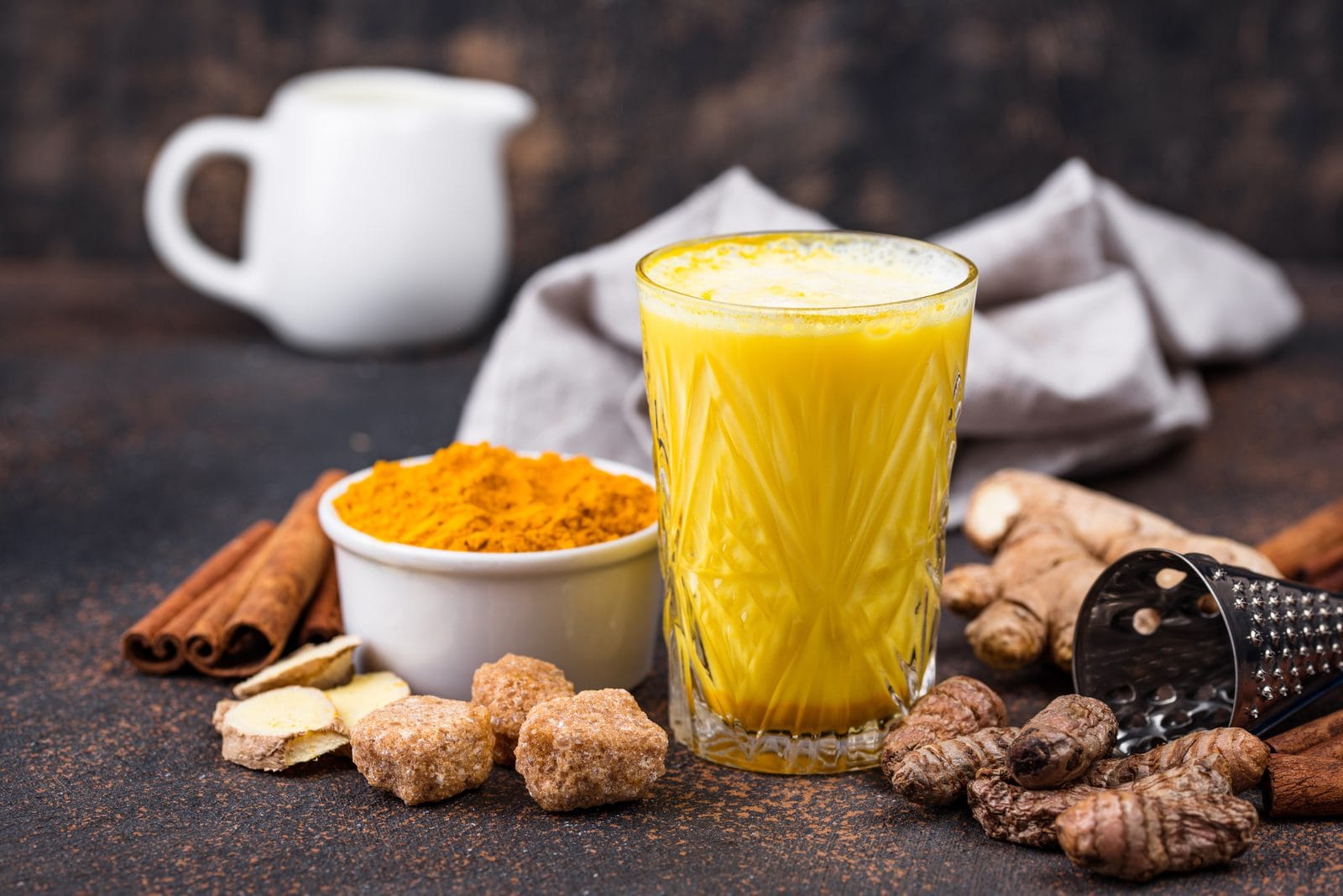 Goldene Milch im Glas neben Kurkuma-Pulver, Zimtstangen und frischem Ingwer