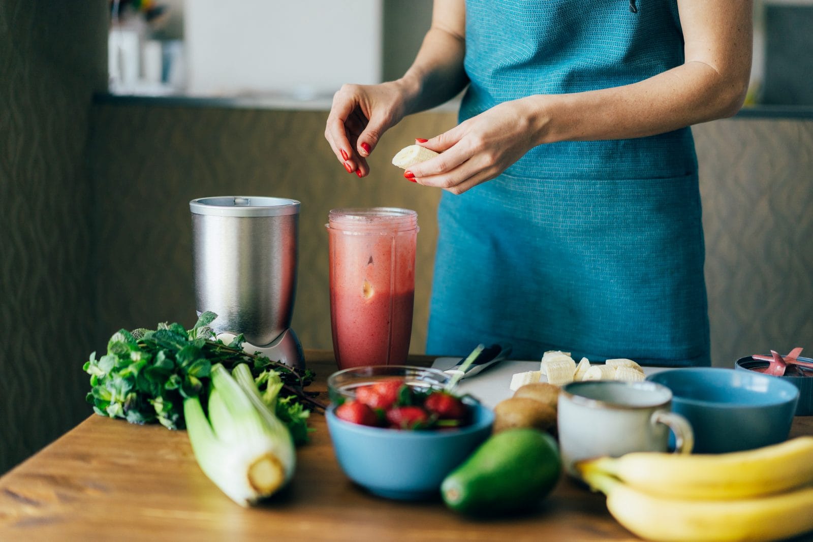 Frau bereitet Smoothie-Zutaten am Mixer vor, frisches Obst und Gemuese auf dem Kuechentisch
