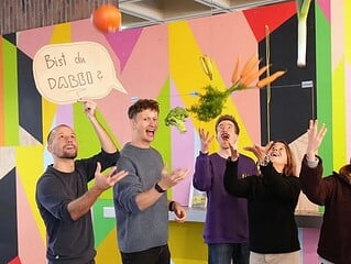 Sechs Menschen werfen Obst und Gemüse in die Luft vor einer bunten Wand, ein Schild fragt Bist du dabei