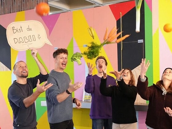 Sechs Menschen werfen Obst und Gemüse in die Luft vor einer bunten Wand, ein Schild fragt Bist du dabei