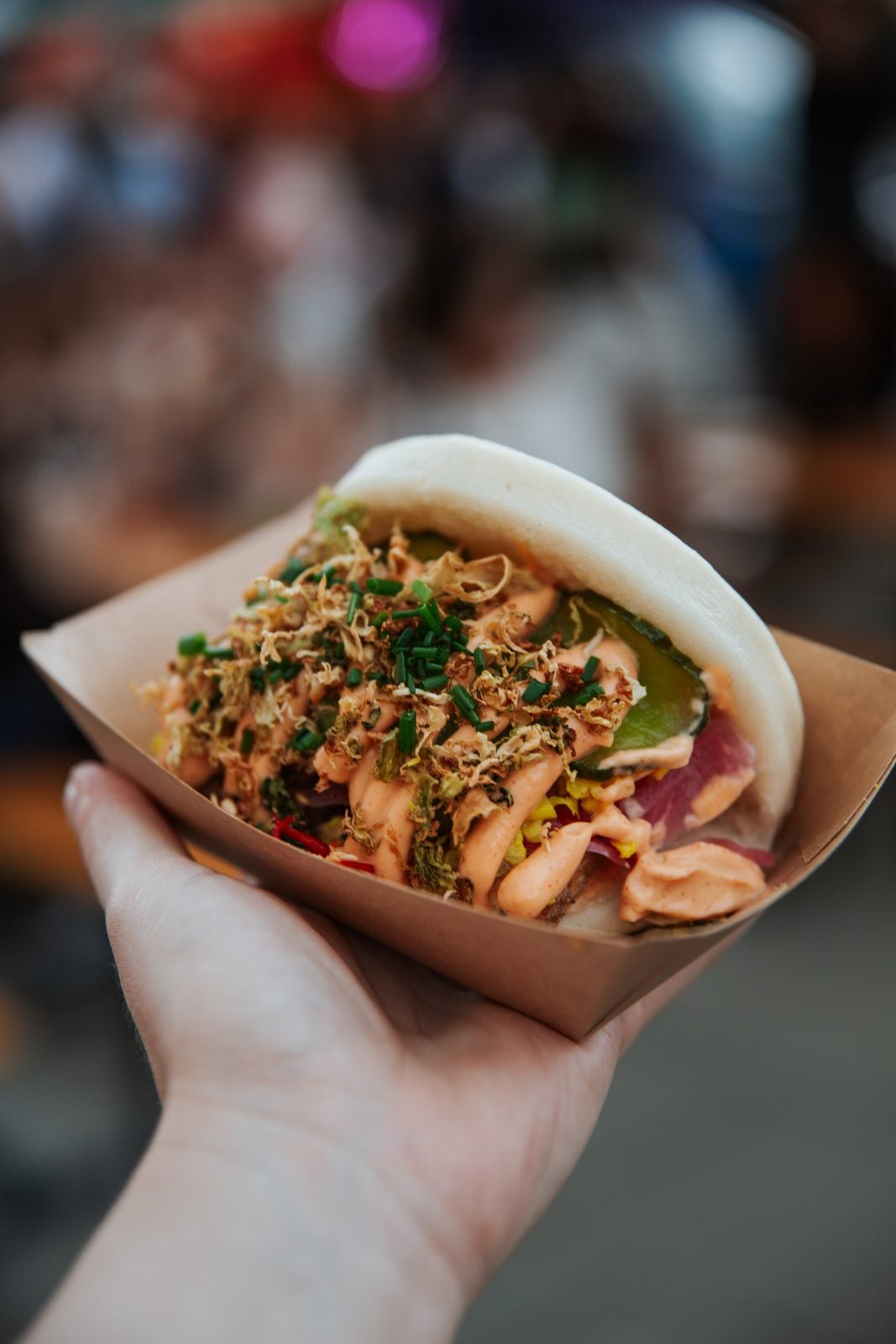 Hand hält einen gefüllten veganen Bao Bun mit Röstzwiebeln und Schnittlauch in einer Pappschale