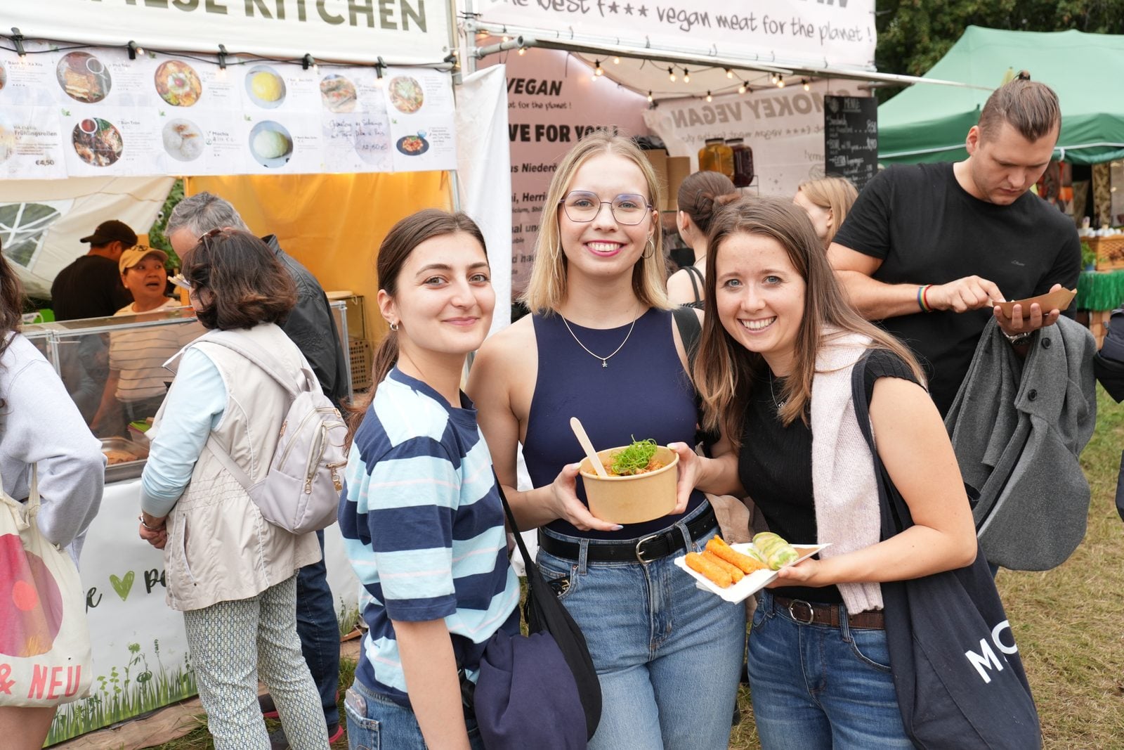 Drei Besucherinnen mit veganem Streetfood in Pappschalen vor einem Essensstand auf der VeganMania Wien