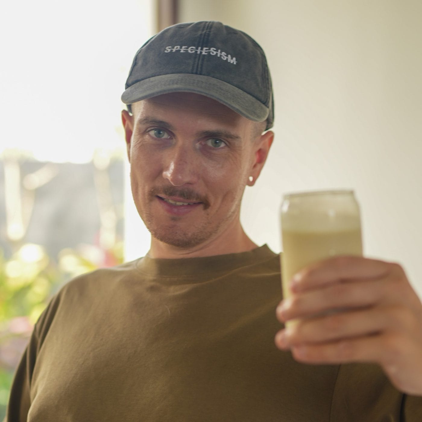 TIV founder Yannick raising a glass of plant milk, wearing a cap with the word speciesism struck through