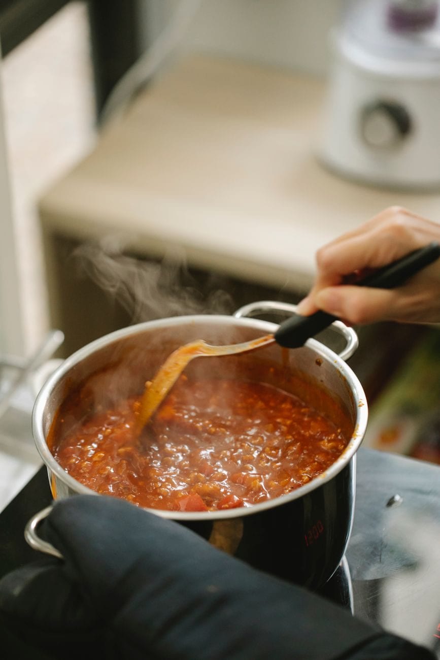 Hand mit Holzlöffel rührt eine rote Tomatensauce in einem Topf auf dem Herd