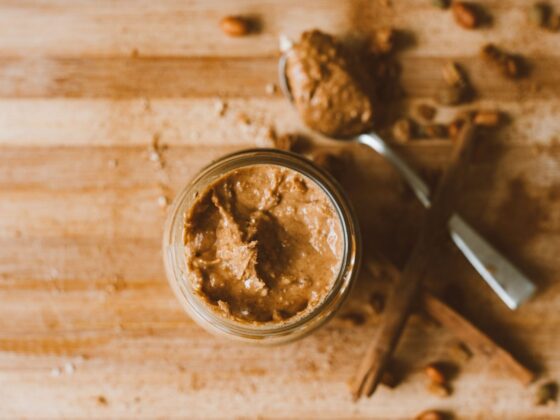 Offenes Glas mit stückiger Erdnussbutter auf einem Holzbrett, daneben ein Löffel, Zimtstangen und Erdnüsse