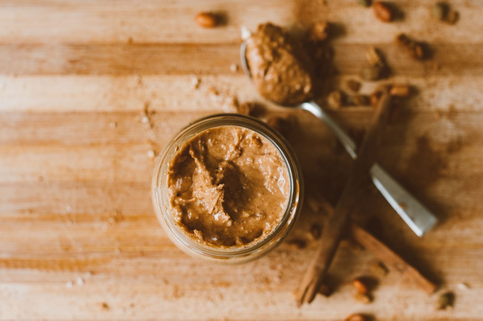 Offenes Glas mit stückiger Erdnussbutter auf einem Holzbrett, daneben ein Löffel, Zimtstangen und Erdnüsse