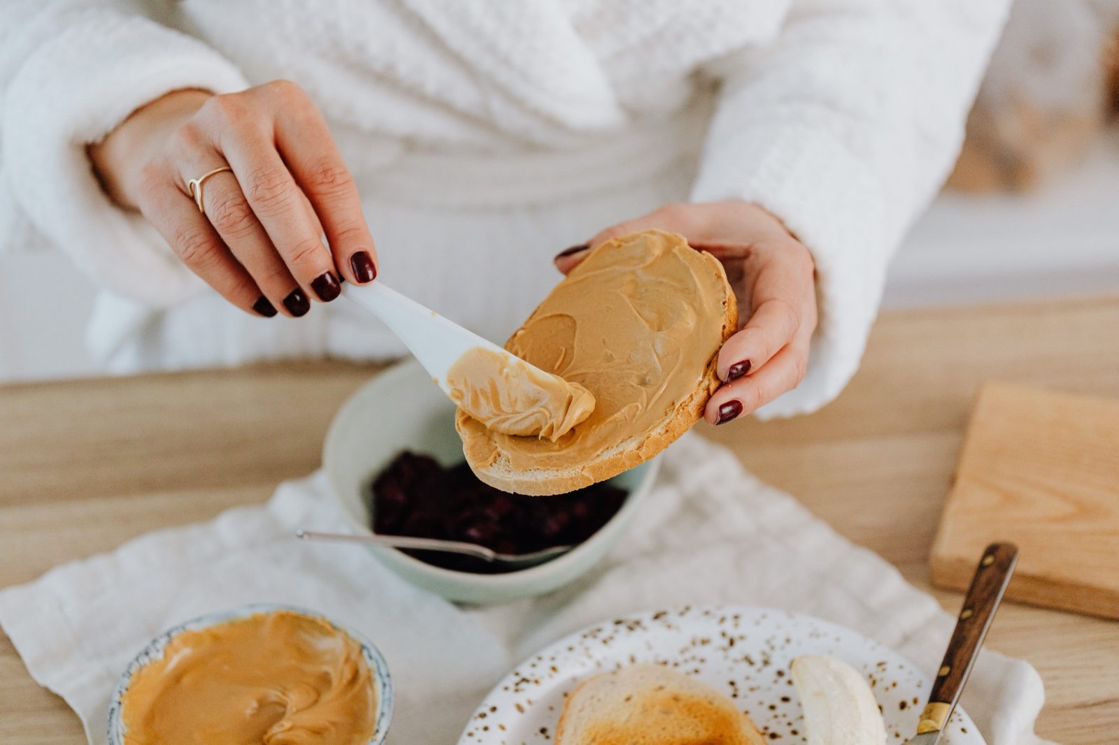 A hand spreading creamy peanut butter on a slice of toast, with berries and half a banana on the table