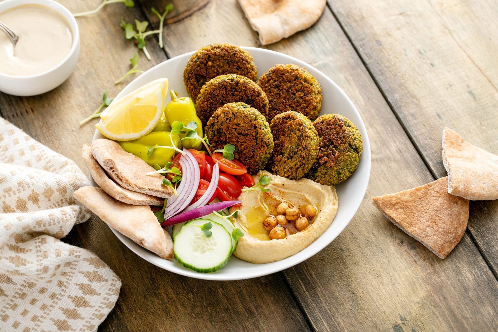 Bowl mit gebackenen Falafeln, Hummus, Gurke, Tomate, roter Zwiebel und Fladenbrot auf einem Holztisch