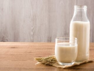 Hafermilch in Glas und Flasche auf einem Holztisch mit Haferähren