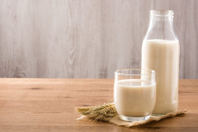 Hafermilch in Glas und Flasche auf einem Holztisch mit Haferähren