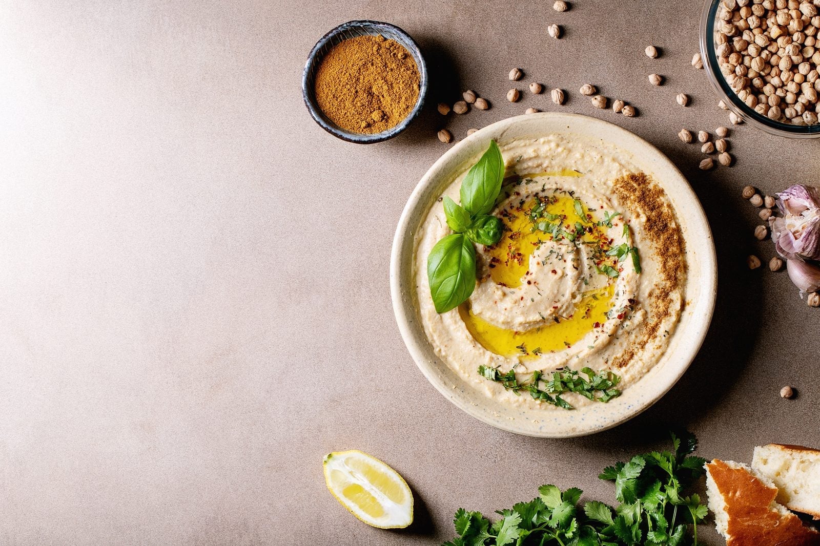 Hummus in einer flachen Schale mit Olivenöl, Basilikum und Kichererbsen, daneben Zitrone und Petersilie