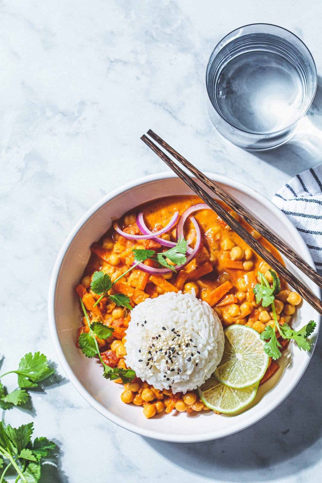Kichererbsen-Curry mit Reis, Limette und Koriander in einer weißen Schale auf Marmor, daneben Stäbchen und ein Glas Wasser