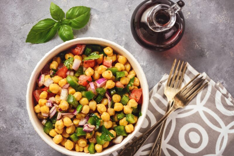 Kichererbsensalat mit grüner Paprika, roter Zwiebel und Basilikum in einer Schüssel, daneben Gabel und Öl