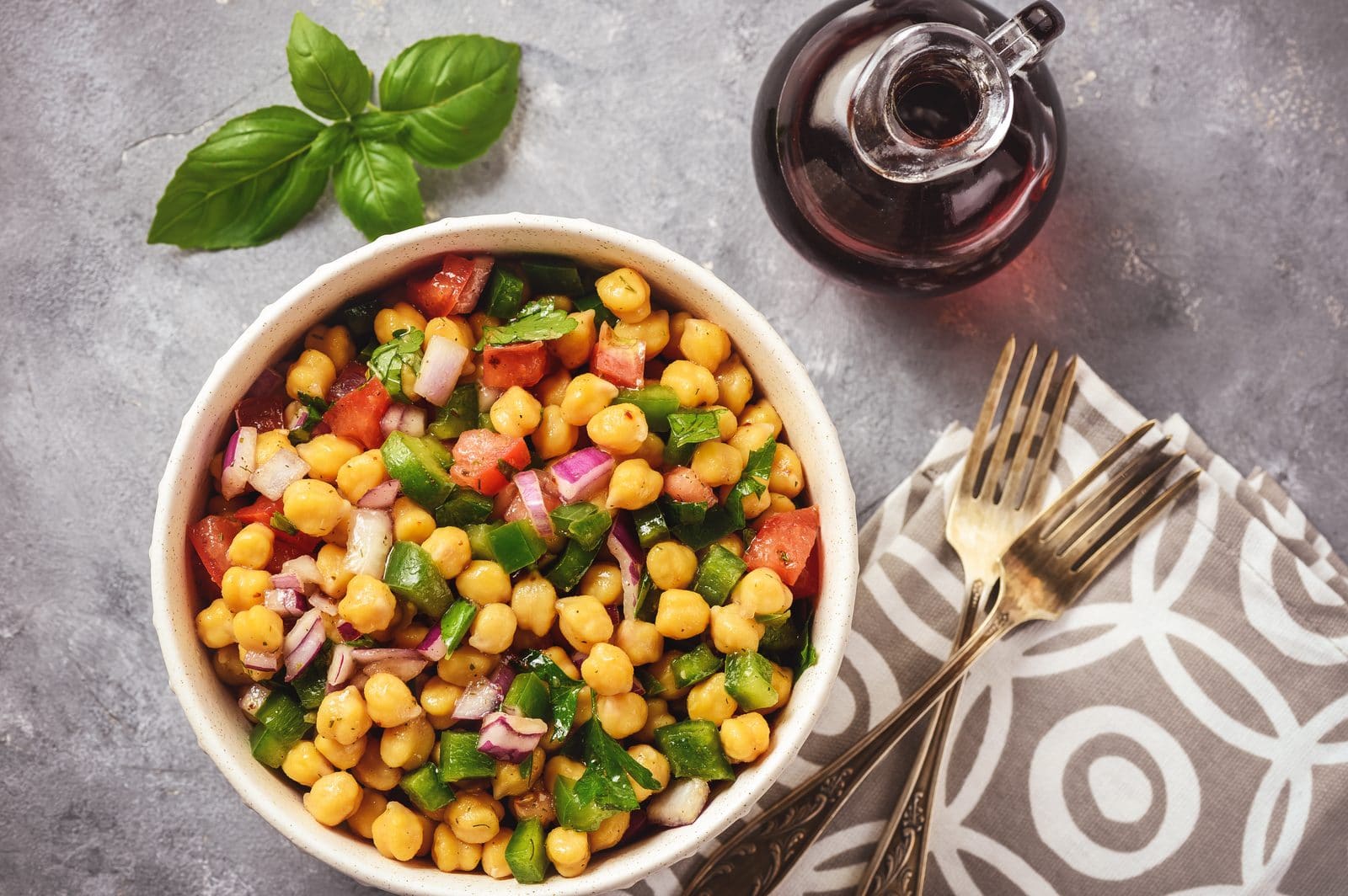Kichererbsensalat mit grüner Paprika, roter Zwiebel und Basilikum in einer Schüssel, daneben Gabel und Öl