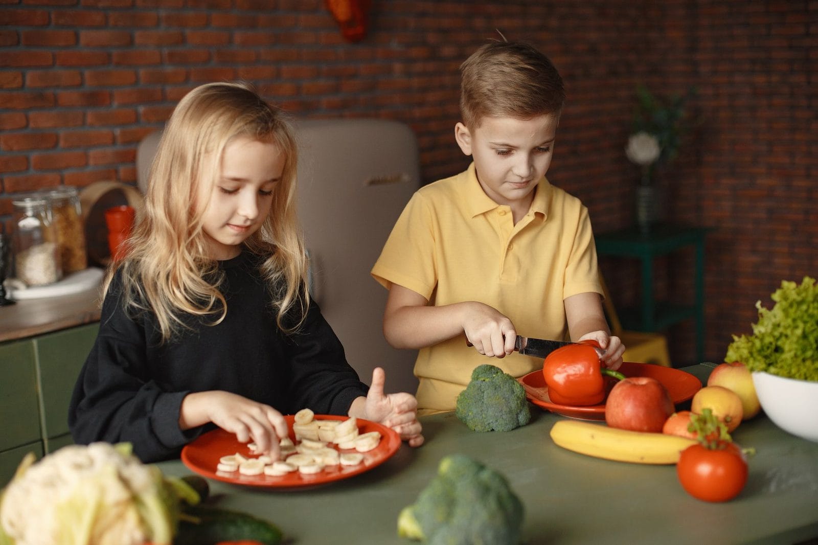 Zwei Kinder schneiden an einem Tisch Obst und Gemüse, daneben liegen Brokkoli, Tomaten und Bananen