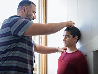 Ein Vater misst die Körpergröße seines Sohnes an einem Türrahmen und markiert sie mit einem Stift