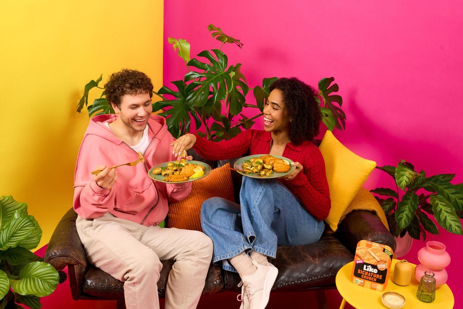 Zwei junge Menschen sitzen lachend auf einem Sofa und essen pflanzliche Schnitzel mit Kartoffeln