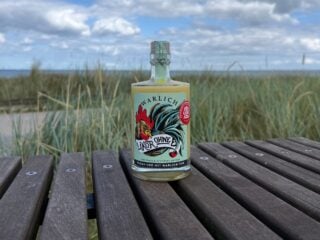 Flasche Likör ohne Ei von Warlich in der zensierten Edition auf einer Holzbank vor Dünengras und blauem Himmel