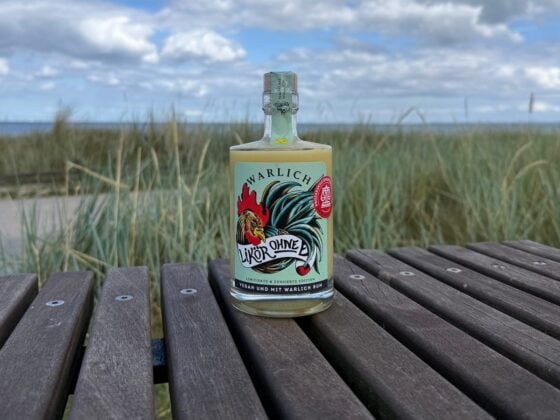 Flasche Likör ohne Ei von Warlich in der zensierten Edition auf einer Holzbank vor Dünengras und blauem Himmel