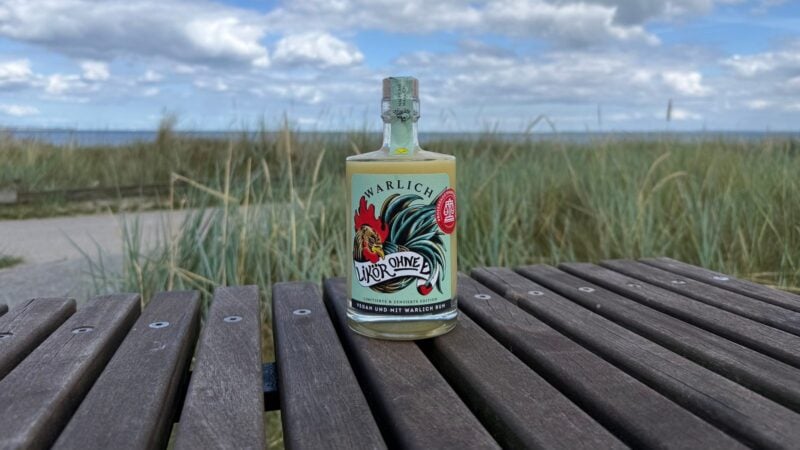 Flasche Likör ohne Ei von Warlich in der zensierten Edition auf einer Holzbank vor Dünengras und blauem Himmel