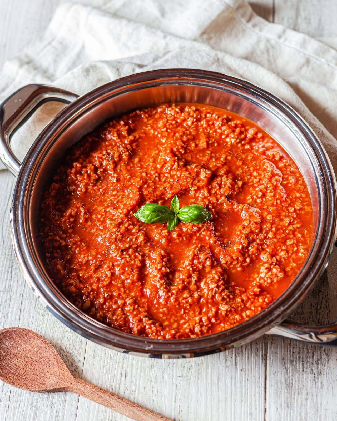 Vegane Bolognese-Sauce in einem Emailletopf von oben, daneben Basilikum und Holzlöffel