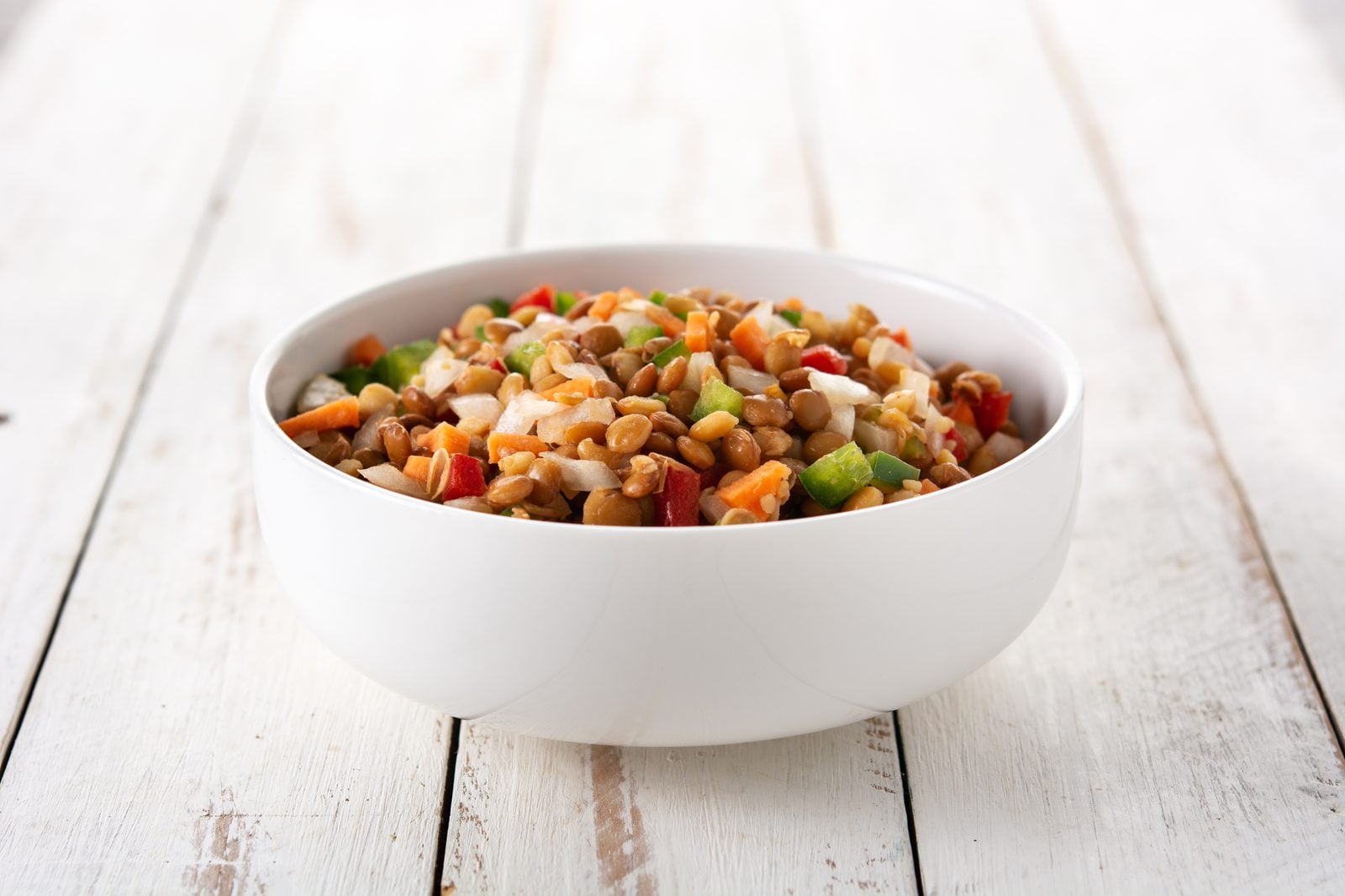 Linsensalat mit Paprikawürfeln, Karotte und Zwiebel in einer weißen Schüssel auf hellem Holztisch