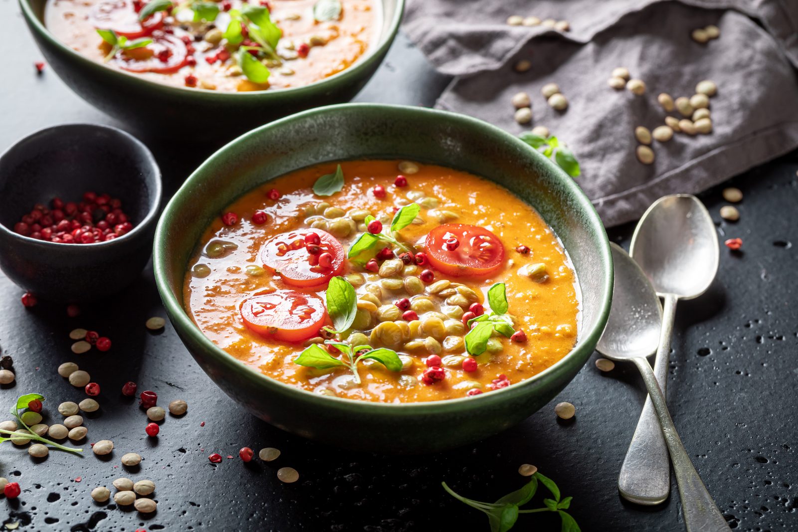 Linsensuppe mit Paprika und Erbsen in einer grünen Schale, daneben rohe Linsen und Hülsenfrüchte