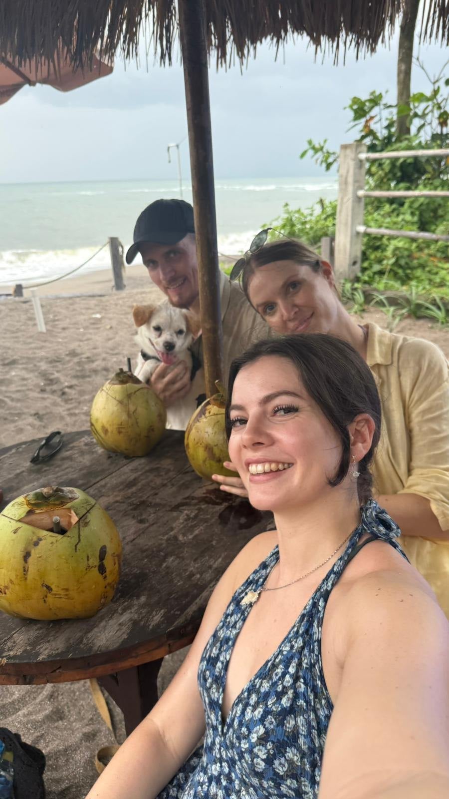 Drei Personen sitzen mit frischen Kokosnüssen an einem Holztisch unter einem Strohschirm am Strand auf Bali