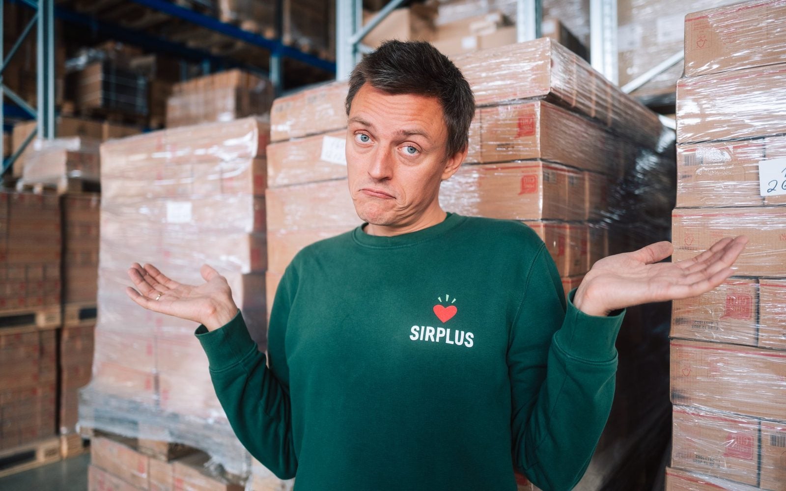 Mann im grünen SIRPLUS-Pullover zuckt vor Paletten mit Kartons fragend mit den Schultern