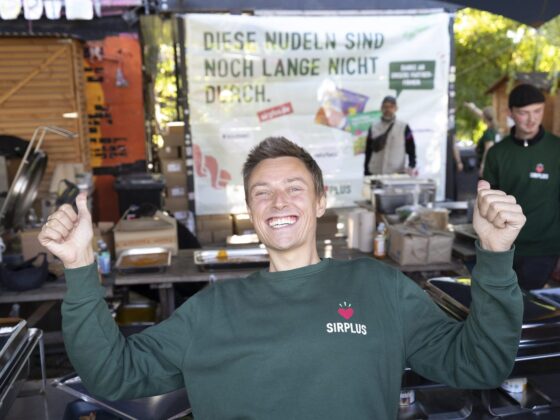 Mann im grünen SIRPLUS-Pullover jubelt mit erhobenen Armen vor vielen Besucher:innen am Holzmarkt