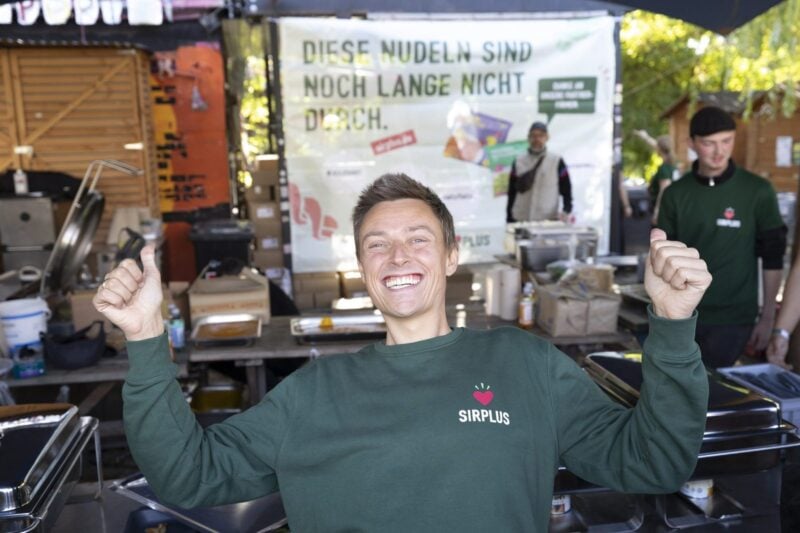 Mann im grünen SIRPLUS-Pullover jubelt mit erhobenen Armen vor vielen Besucher:innen am Holzmarkt